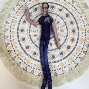 May include: A handmade doll with a printed face, wearing a dark blue t-shirt and blue jeans. The doll is posed on a round, patterned mat with a fringed edge. The mat features geometric shapes in blue, yellow, and white.