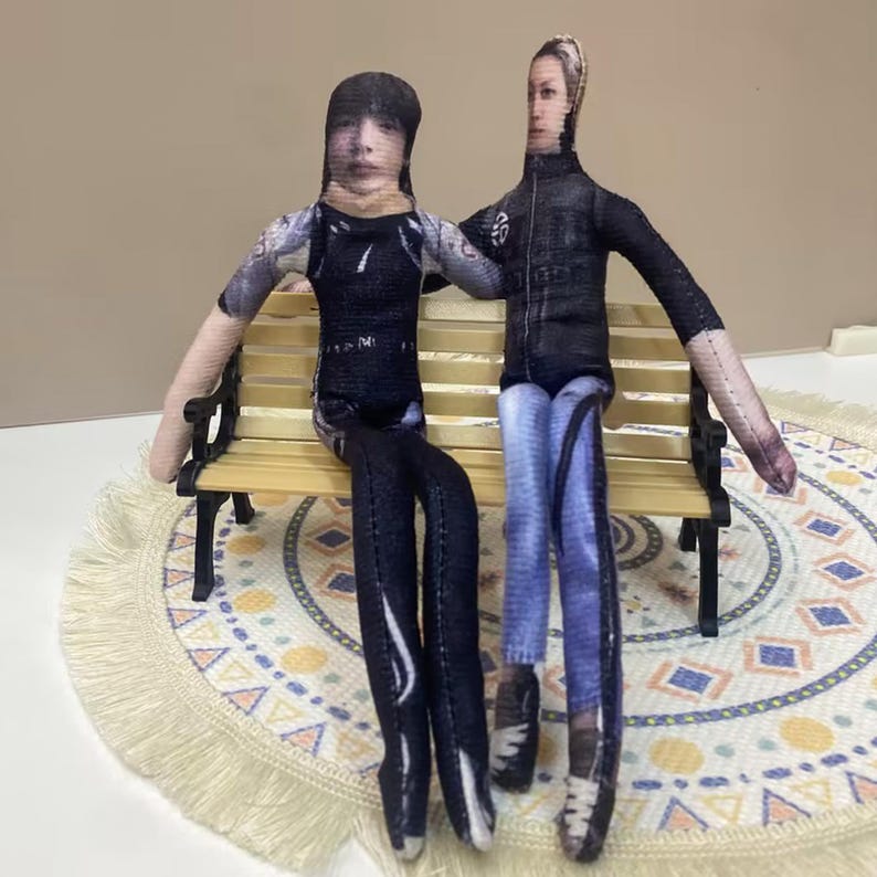 May include: Two fabric dolls sit on a miniature park bench. One doll wears a black top and pants, the other a black jacket and blue jeans. The dolls have printed faces and are posed as if they are embracing. The bench is made of wood and has black legs.