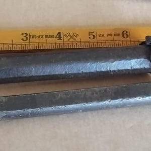 May include: Two dark grey, hand-forged metal chisels with textured, hammered surfaces and flat, beveled edges. The tools are positioned parallel to a yellow wooden ruler, showing the top chisel is approximately 16.5 cm in length.