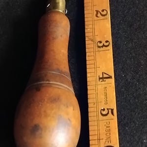 Könnte beinhalten: Ein Werkzeug mit einem Vintage-Holzgriff, einer Messingzwinge und einer Montageplatte, platziert neben einem gelben Holzlineal, das eine Länge von etwa 16,5 cm anzeigt. Das Werkzeug hat einen glatten, abgerundeten, braunen Holzgriff mit Ringen.