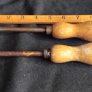 May include: Two vintage hand tools with wooden handles and metal shafts, each featuring a flat metal head. One tool has a circular head, the other a triangular head. A yellow wooden ruler is positioned above, showing a length of 11 inches.