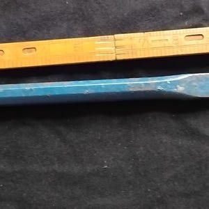 May include: A blue metal cold chisel with a hexagonal shaft and a flat, beveled cutting edge, positioned alongside a yellow wooden folding ruler that shows a measurement of approximately 8 inches for the length of the tool.