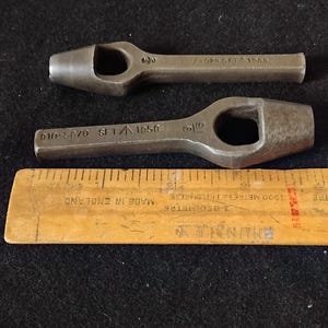 Könnte beinhalten: Zwei Vintage-Metalllochstanzen mit rechteckigen Öffnungen, neben einem Holzlineal. Die Lochstanzen haben eingestempelte Markierungen, einschließlich Zahlen und Buchstaben. Das Lineal, mit der Aufschrift "MADE IN ENGLAND", dient als Größenreferenz.