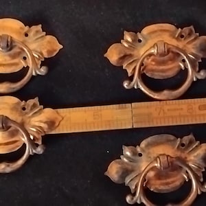 Op de afbeelding: Vier messing ladehandgrepen met versierde, bladvormige achterplaten en ronde ringgrepen. Een houten liniaal is over de handgrepen geplaatst en toont een breedte van ongeveer 9 centimeter voor elk van de afzonderlijke handgrepen.