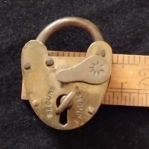 May include: A brass heart-shaped padlock with a metal keyhole cover featuring a star engraving. The word SECURE is embossed on the front. A wooden ruler is positioned behind the lock, showing a width of approximately 3.8 cm for the body.