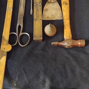 May include: A collection of vintage tools including a yellow folding wooden ruler, metal scissors, a small screwdriver with a black handle, a brass spirit level, a metal putty knife with a wooden handle, a small wooden top, and a ball-peen hammer.