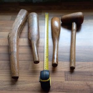 May include: A collection of antique wooden tools, including mallets and pestle-like objects, arranged on a wooden surface. A yellow measuring tape is placed between the tools. The tools vary in shape and size, with a natural wood finish.