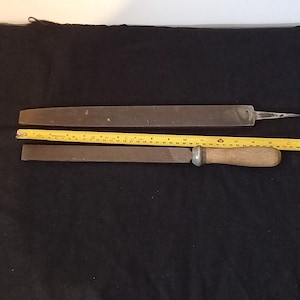 May include: Two metal files and a yellow measuring tape are displayed against a dark background. One file is long and thin, while the other is shorter with a wooden handle. The measuring tape shows inches.