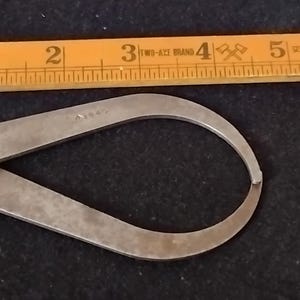 May include: A vintage metal caliper tool with a curved design, next to a wooden ruler. The caliper is silver-toned and has a hinged joint. The ruler is yellow with black markings, showing measurements in inches.