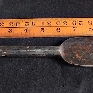 May include: A metal cold chisel with a hexagonal shaft and a flat, wide blade showing signs of oxidation. The tool is positioned alongside a wooden folding ruler, indicating a total length of approximately 7.5 inches for the metal chisel.