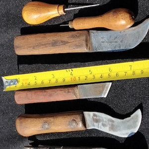 Pode incluir: Uma coleção de ferramentas de marcenaria vintage, incluindo furadores, facas e alicates, dispostas sobre uma superfície escura. As ferramentas apresentam cabos de madeira e lâminas de metal, com uma fita métrica amarela para escala. As ferramentas são provavelmente usadas para moldar e cortar madeira.