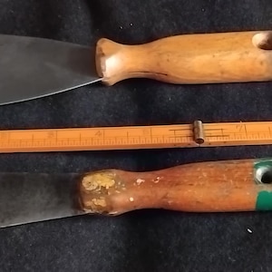 May include: Two metal putty knives with wooden handles positioned next to a wooden ruler. The top knife has a wide, flat blade, while the bottom knife has a pointed blade. The ruler shows measurements in centimetres, indicating a length of 20 centimetres.