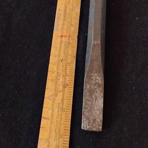 May include: A vintage metal chisel and a wooden ruler. The chisel has a rectangular body and a flat, beveled edge. The wooden ruler has black markings and red accents. The items are on a dark background.