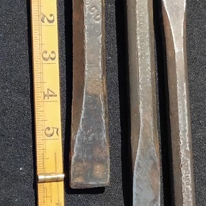 May include: Three weathered metal cold chisels with textured, dark grey surfaces and flared heads, positioned next to a yellow folding ruler that shows the tools are approximately 16.5 cm in length. The metal surfaces show signs of wear.