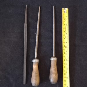 Może przedstawiać: Trzy metalowe pilniki z drewnianymi uchwytami i żółta linijka. Pilniki mają różne kształty, w tym pilnik płaski i dwa okrągłe. Linijka pokazuje pomiary w centymetrach.