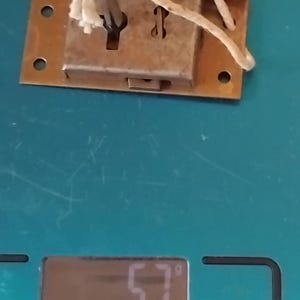 May include: A rectangular brass-coloured metal lock plate with a key inserted into the centre keyhole. A piece of off-white string is tied through the key handle and the lock plate. The lock is resting on a digital scale displaying a weight of 57 grams.