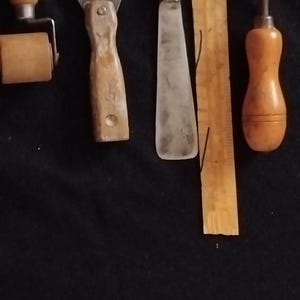 Könnte beinhalten: Eine Sammlung alter Werkzeuge, bestehend aus einer Holzrolle, einem Metallspachtel mit Holzgriff, einem langen Metallspatel mit dunklem Holzgriff, einem Holzlineal und einem Metallwerkzeug mit Holzgriff, angeordnet auf einer schwarzen Fläche.