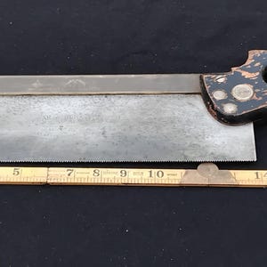 May include: An antique hand saw with a dark handle and a metal blade. The blade is approximately 30 cm long. A wooden ruler is placed next to the saw for measurement. The saw has a vintage, worn appearance.