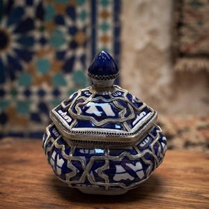 Peut inclure: Un pot hexagonal avec couvercle présentant un motif géométrique bleu profond et blanc. Le corps en céramique est recouvert d'une superposition décorative en métal argenté suivant les contours du couvercle et de la base, avec un fleuron pointu.