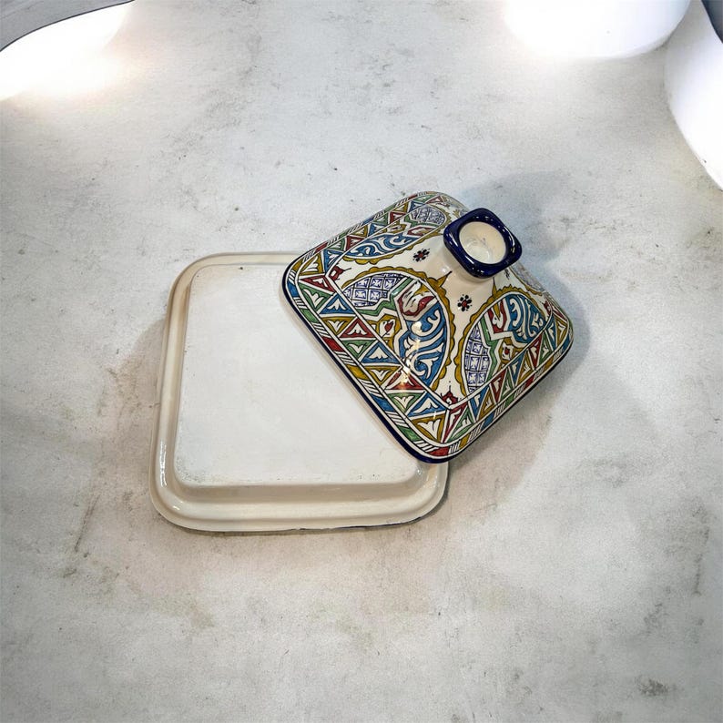 May include: A square, off-white ceramic tagine dish with a matching lid. The lid features a colorful, intricate geometric pattern in red, blue, green, and yellow. The dish is open, revealing the interior.