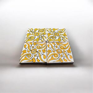 Puede incluir: Un conjunto de cuatro azulejos de cerámica cuadrados dispuestos en cuadrícula, con fondo blanco y motivos florales amarillos pintados a mano junto a patrones de enredaderas negras. Cada azulejo muestra un diseño simétrico de flores y hojas.