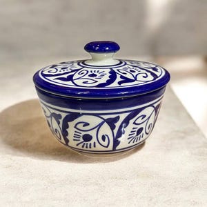Op de afbeelding: Een kleine, ronde keramische container met een deksel. De container is wit met een blauw bloemmotief en een blauwe rand. Het deksel heeft een blauwe knop. De container wordt waarschijnlijk gebruikt voor het opbergen van kleine items.