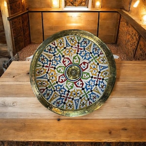 May include: A round decorative tray with a gold-coloured rim and a colourful, intricate pattern. The pattern features geometric shapes in blue, green, red, yellow, and white. The tray is placed on a wooden surface.