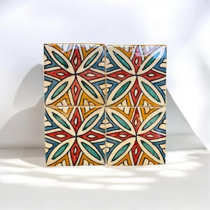 May include: A set of four square ceramic tiles arranged in a larger square formation. Each tile features a symmetrical geometric pattern with a central star motif in red, teal, and yellow, outlined in black against a cream-coloured background.