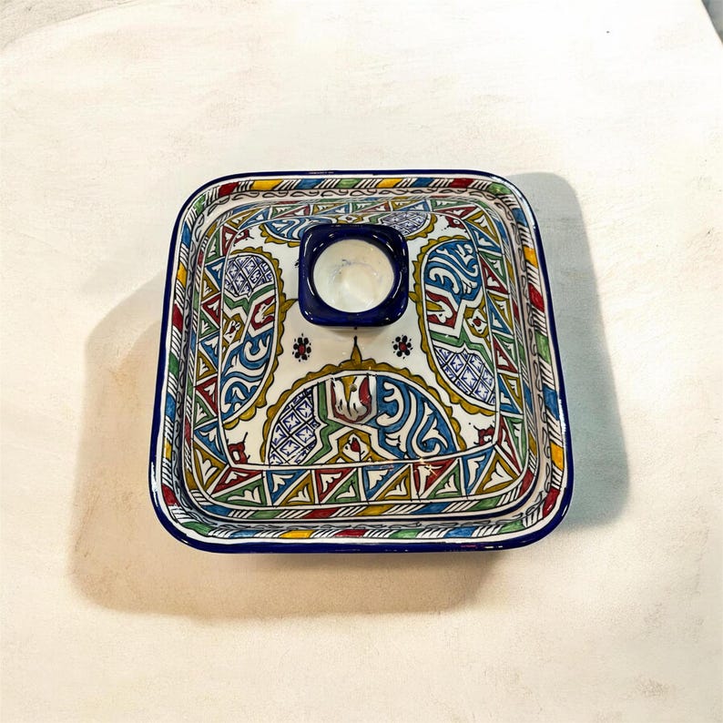 May include: Square ceramic dish with a lid, featuring a vibrant, intricate pattern in blue, yellow, red, and green. The dish has a dark blue rim and a small, circular opening on the lid. The design is geometric and floral.