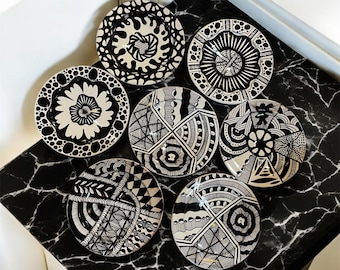 May include: Seven decorative plates with black and white patterns are arranged on a black and white marble surface. Each plate features a unique design, including floral, geometric, and abstract motifs. The plates are round and appear to be ceramic.