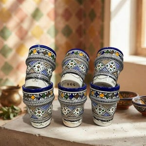 Peut inclure: Un ensemble de six verres à thé en céramique ornés de motifs géométriques complexes en bleu, vert, jaune et blanc, agrémentés de bandes en filigrane métallique argenté. Chaque verre porte l'inscription FES sur la base, disposés en deux piles.