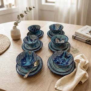 Peut inclure: Un ensemble de six bols en céramique avec soucoupes assorties, ornés de motifs floraux complexes en bleu et blanc. Les bols sont disposés en deux rangées sur une surface en bois, avec une cuillère en argent et une serviette en tissu beige.