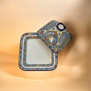 May include: Square ceramic serving dish with a lid. The dish and lid are white with colorful geometric patterns in red, blue, green, and yellow. The lid has a small blue handle. The dish is open, revealing the interior.