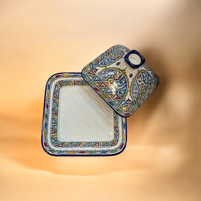 May include: Square ceramic serving dish with a lid. The dish and lid are white with colorful geometric patterns in red, blue, green, and yellow. The lid has a small blue handle. The dish is open, revealing the interior.
