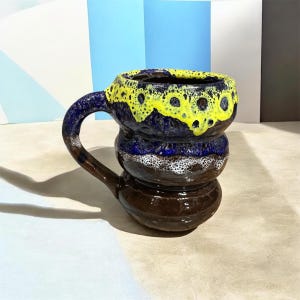 Peut inclure: Une tasse en céramique avec un corps arrondi à plusieurs niveaux et une anse incurvée. L'émail présente un motif de gouttes jaune vif sur le bord, passant par une bande bleu profond, une section centrale blanche tachetée et une base marron.