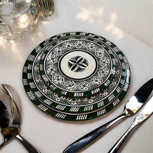 May include: Three decorative ceramic plates with a green and white geometric pattern. The plates are stacked. Silverware and a glass pitcher are in the background. The plates have a central floral design.