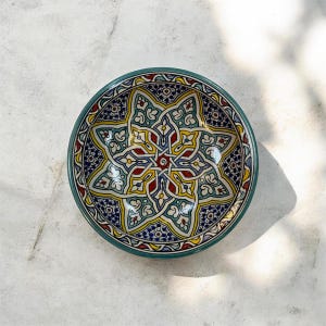 Peut inclure: Un bol rond en céramique orné d'un motif complexe d'étoile géométrique peint à la main dans des tons vifs de bleu sarcelle, jaune, rouge et bleu foncé. Le design intérieur présente des formes entrelacées et des motifs floraux, avec un bord bleu sarcelle.