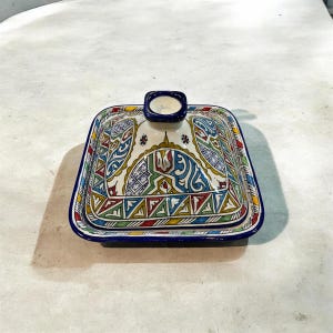 May include: A square ceramic dish with a lid, featuring a vibrant, intricate pattern in blue, red, green, and yellow. The dish has a blue rim and a small, rectangular handle on the lid. The design is likely hand-painted.