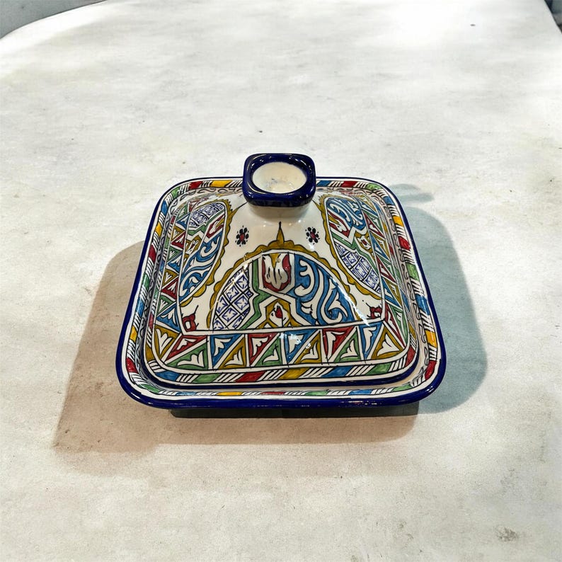 May include: A square ceramic dish with a lid, featuring a vibrant, intricate pattern in blue, red, green, and yellow. The dish has a blue rim and a small, rectangular handle on the lid. The design is likely hand-painted.