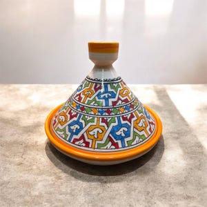 May include: A colorful ceramic tagine pot with a conical lid and a wide, shallow base. The pot features a vibrant geometric pattern in red, blue, green, and yellow against a white background. The rim and lid handle are orange.