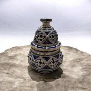 Peut inclure: Un pot décoratif avec couvercle, doté d'une base arrondie et d'un couvercle assorti. La surface est ornée d'un motif géométrique bleu foncé et blanc, rehaussé par des bordures en métal argenté, des détails en corde et des cristaux clairs.