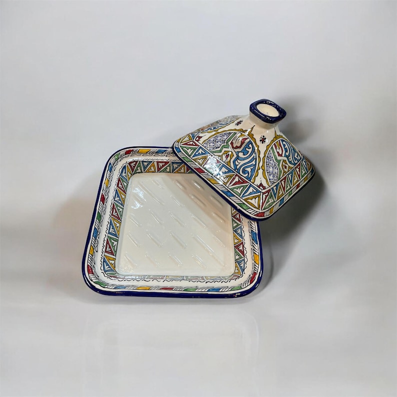May include: A square, ceramic tagine dish with a lid. The dish is white with a blue rim and colorful geometric patterns. The lid features a handle and similar decorative designs in blue, green, red, and yellow.