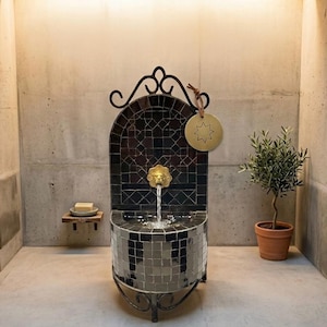 May include: A wall-mounted fountain featuring dark, square mosaic tiles and a central brass lion head spout. The fountain has a curved metal frame with scrollwork at the top and base. A circular brass tag with a star engraving hangs from the top.