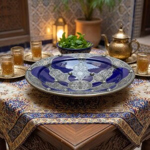 May include: A blue and silver decorative serving platter with an ornate design sits on a patterned tablecloth. The table is set with glasses, small gold trays, and a teapot, suggesting a tea service. The scene is set outdoors.