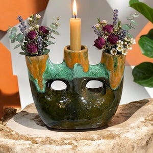 Peut inclure: Un bougeoir en céramique fait à la main avec un glaçage vert, orange et marron. Il comporte un porte-bougie central avec une bougie allumée et deux vases latéraux remplis de fleurs séchées. Le bougeoir est posé sur une surface en pierre.