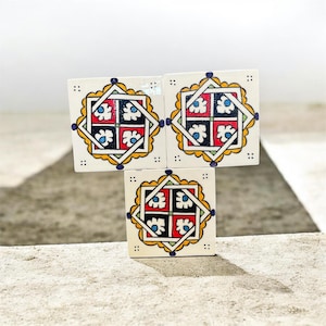 Peut inclure: Trois carreaux de céramique carrés présentant un motif géométrique peint à la main. Chaque carreau a un fond blanc avec un carré central divisé en quatre quadrants rouges et noirs, ornés de motifs floraux bleus, encadrés de lignes jaunes et blanches.