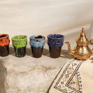 Peut inclure: Ensemble de verres à thé marocains aux bords colorés orange, vert, bleu et marine. Une théière dorée, un vase blanc et des dattes sont disposés sur une surface en pierre avec un tapis à motifs, créant une scène chaleureuse.