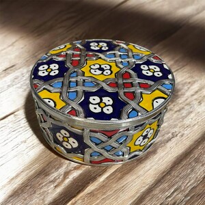 Puede incluir: Una caja redonda y decorativa con un marco de metal plateado y una tapa estampada y colorida. La tapa presenta un diseño geométrico con detalles en amarillo, azul, rojo y blanco. La caja está sobre una superficie de madera.