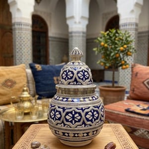 Puede incluir: Un jarrón decorativo de cerámica con tapa, con un patrón geométrico azul y blanco y detalles plateados. El jarrón se exhibe sobre una mesa de madera, mostrando un diseño tradicional. El entorno incluye un patio con cojines y un pequeño árbol.