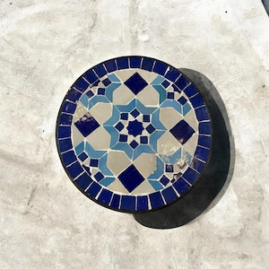 Puede incluir: Un tablero de mesa redondo con un patrón de mosaico de azulejos azul oscuro, azul claro y beige. El diseño consiste en una estrella central rodeada de formas geométricas, con un borde azul oscuro sólido que rodea toda la superficie circular.